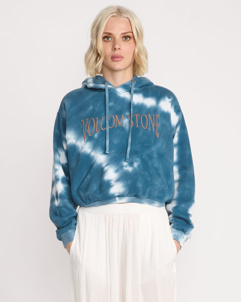 A woman with blonde, shoulder-length hair wears the Volcom Women's New Tie Dye Hoodie featuring blue and white tones with orange VOLCOM STONE lettering, paired with a white skirt against a plain white background.