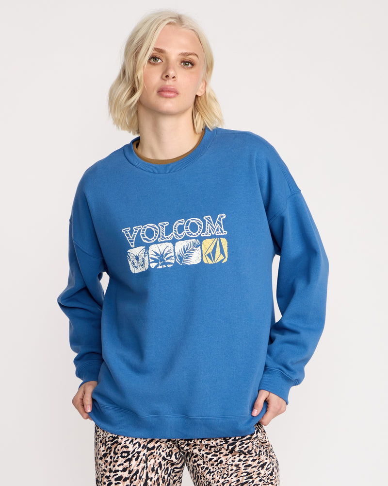 A woman with short blonde hair wears the Volcom Women's Stone Crew sweatshirt in a boyfriend fit, featuring white and yellow graphics. The brushed-back fleece pops against a white background, styled with leopard print pants.