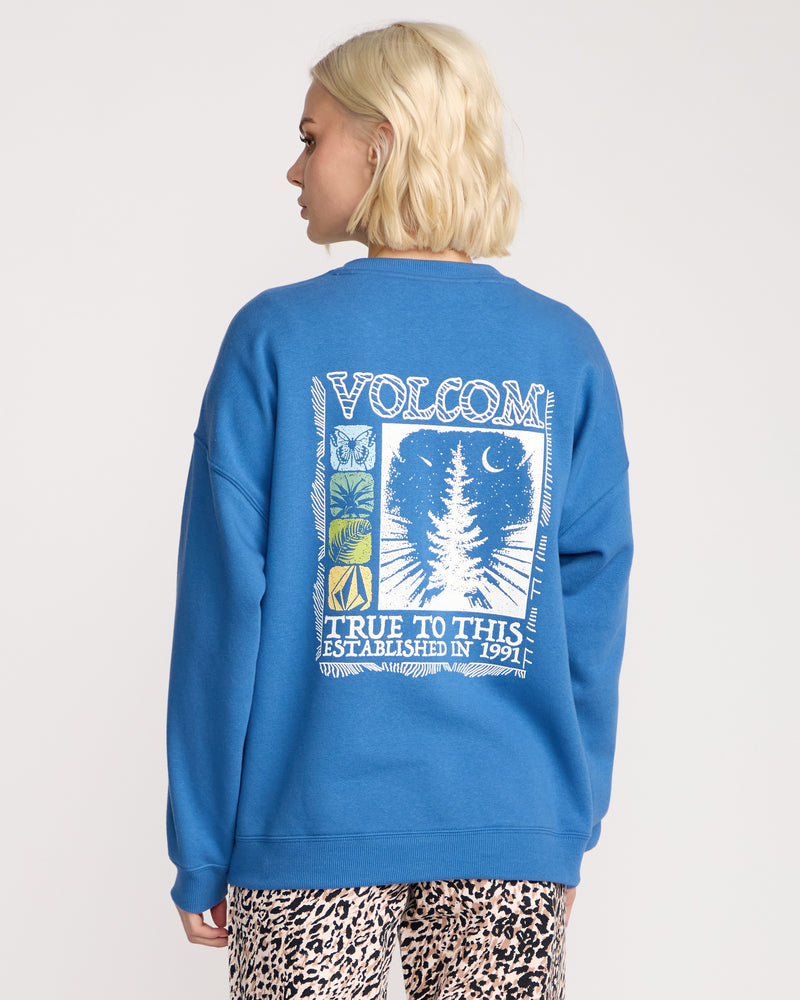A woman with short blonde hair wears the Women's Stone Crew by Volcom, a bright blue cozy boyfriend-fit sweatshirt with tree and crescent moon graphics and "TRUE TO THIS ESTABLISHED IN 1991" on the back, paired with leopard-print pants.