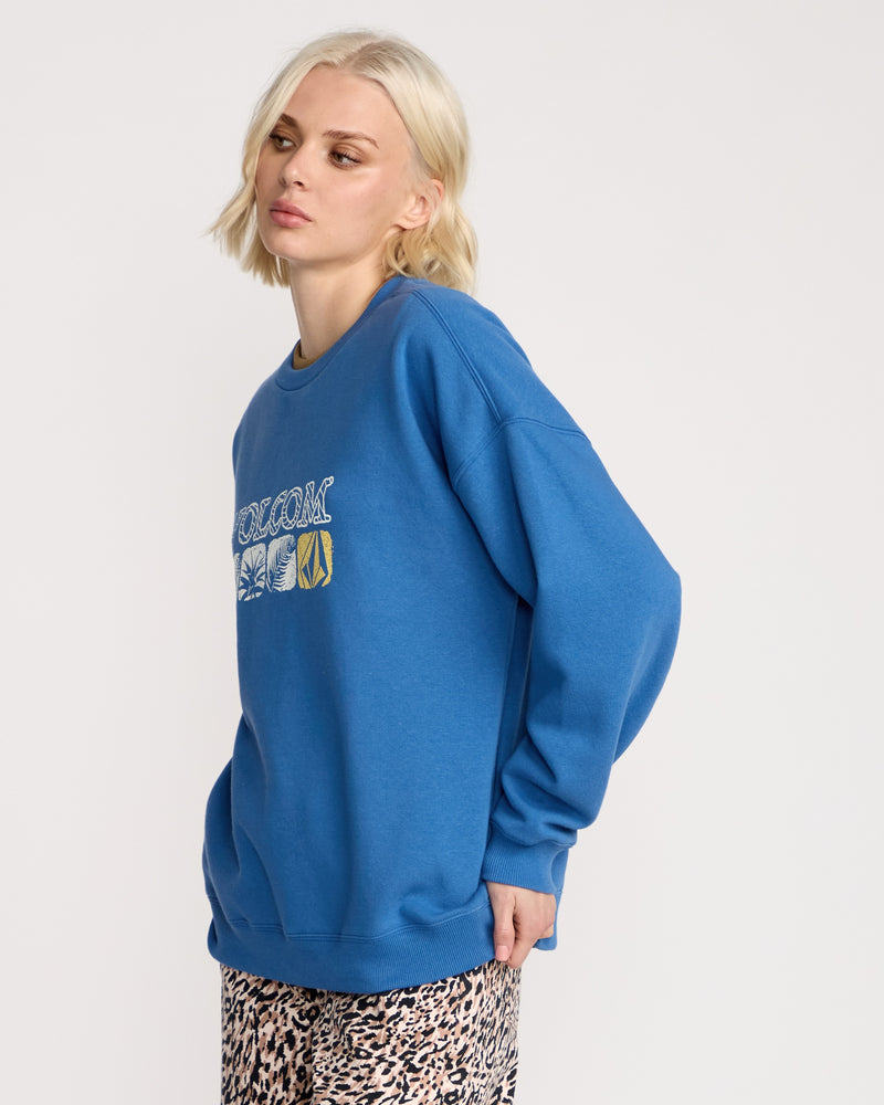 A woman with short blonde hair wears a loose Volcom Women's Stone Crew sweatshirt in brushed-back fleece, featuring graphic designs, along with patterned pants, standing against a plain light background.