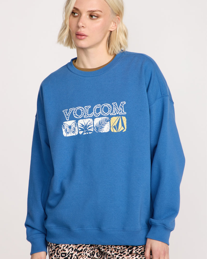A woman with short blonde hair wears the Volcom Women's Stone Crew sweatshirt in blue, featuring white and yellow leaf graphics and a cozy boyfriend fit, paired with leopard print pants against a plain light background.