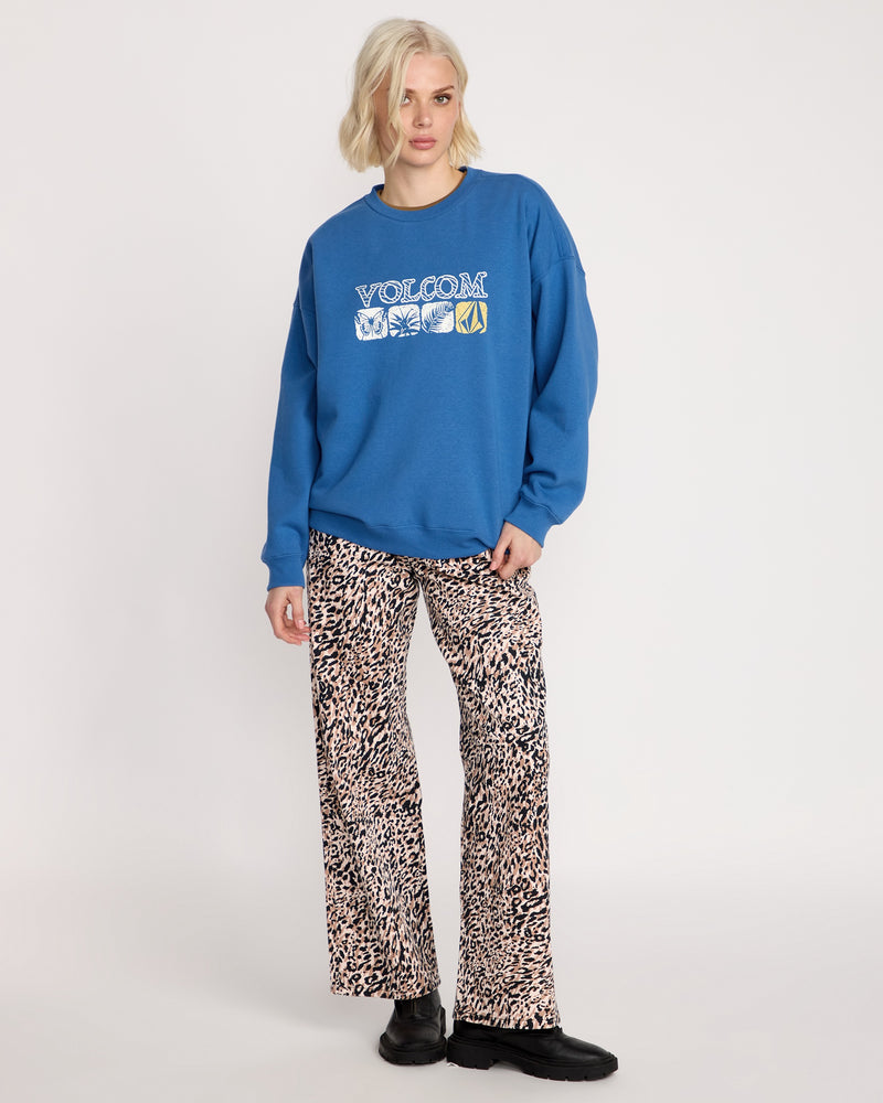 A person with chin-length blonde hair wears a blue Volcom Women's Stone Crew sweatshirt in a boyfriend fit, paired with leopard print pants and black shoes, standing against a plain white background.