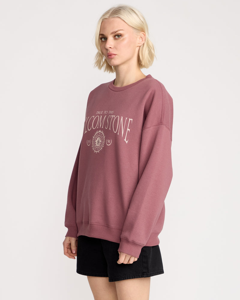 A person with short blonde hair wears the Volcom Women's Stone Crew, a mauve fleece pullover with white text and emblem, paired with black shorts, standing against a plain white background.
