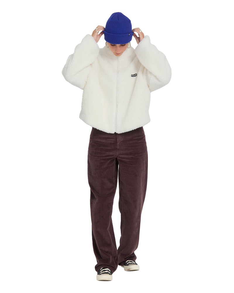 A person wearing an oversized white Volcom Women’s Wylding Out Sherpa zip-up jacket, brown corduroy pants, a blue beanie, and black sneakers.