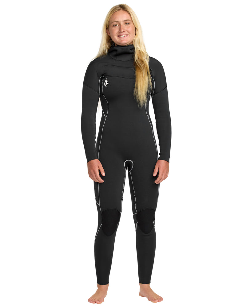 A smiling woman with long blonde hair stands barefoot against a white background, wearing the Volcom Women's 4/3 Modulator Chest Zip Hooded Full Wetsuit in black with white accent lines.