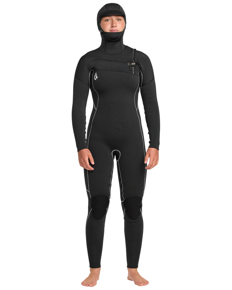 A woman stands barefoot facing forward in the Volcom Women's 4/3 Modulator Chest Zip Hooded Full Wetsuit, featuring white seam lines and reinforced knee pads, with arms relaxed at her sides.