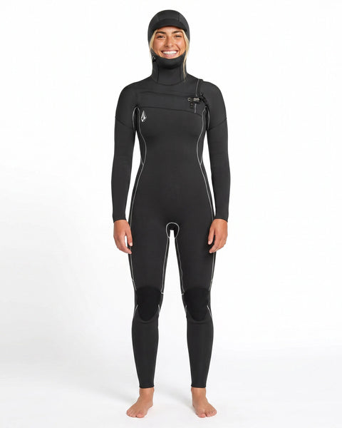 A person stands barefoot and smiling against a plain white background, wearing the Volcom Women's 4/3MM Modulator Chest Zip Hooded Full Wetsuit with white seams.