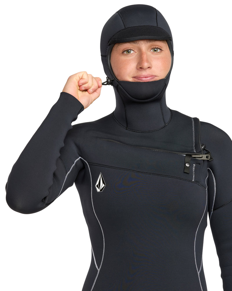 A woman wearing the Volcom Women's 5/4 Modulator Chest Zip Hooded Full Wetsuit with white stitching and a chest logo is pulling up the neck zipper and looking at the camera.