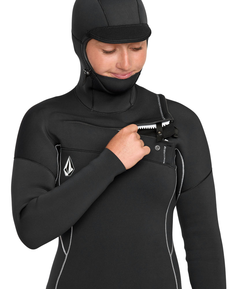 A woman in a Volcom Women's 5/4 Modulator Chest Zip Hooded Full Wetsuit with white stitching and a small chest logo unzips the front, looking down with a slight smile.