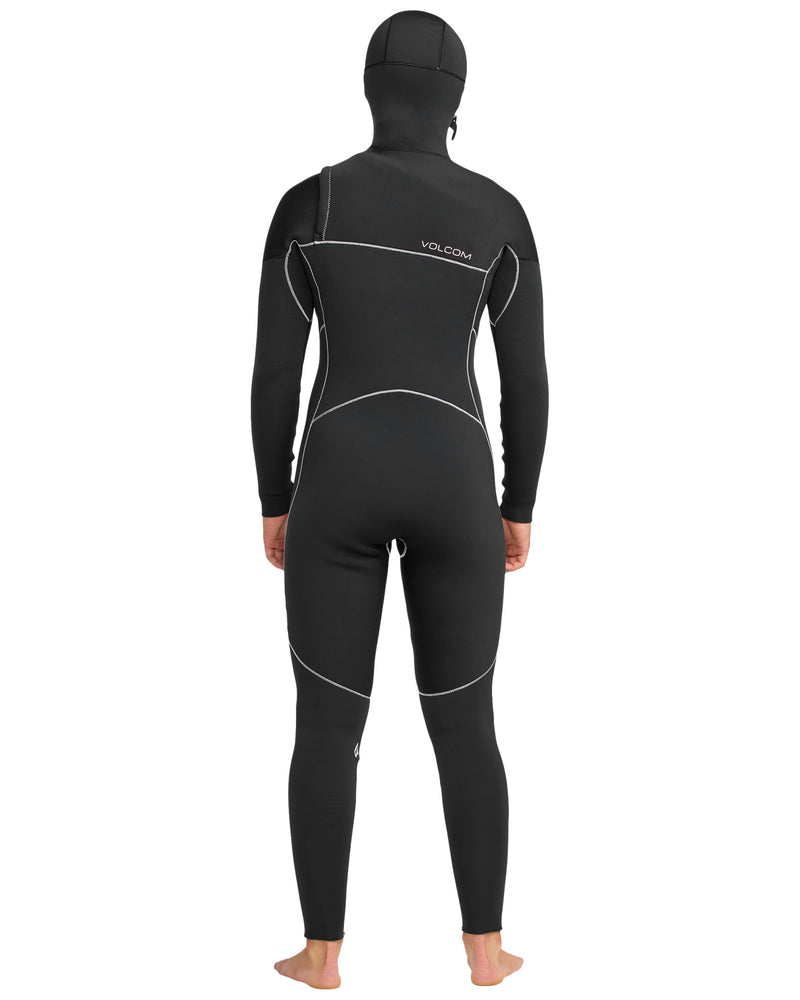 A person stands barefoot, facing away, wearing the Volcom Women's 5/4 Modulator Chest Zip Hooded Full Wetsuit. The snug black wetsuit features white seams, an attached hood, and the Volcom logo on the upper back.