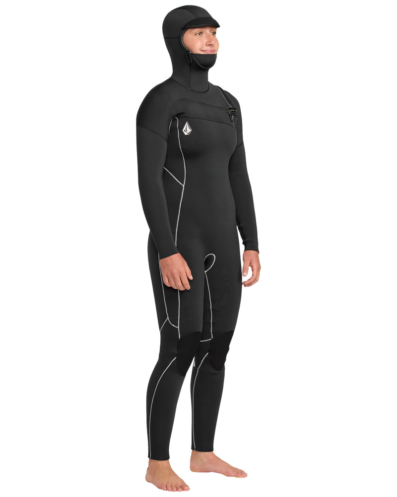 A person is barefoot wearing the Volcom Women's 5/4 Modulator Chest Zip Hooded Full Wetsuit, featuring white seam lines and a chest zipper. The black suit covers the body except for the face, hands, and feet.