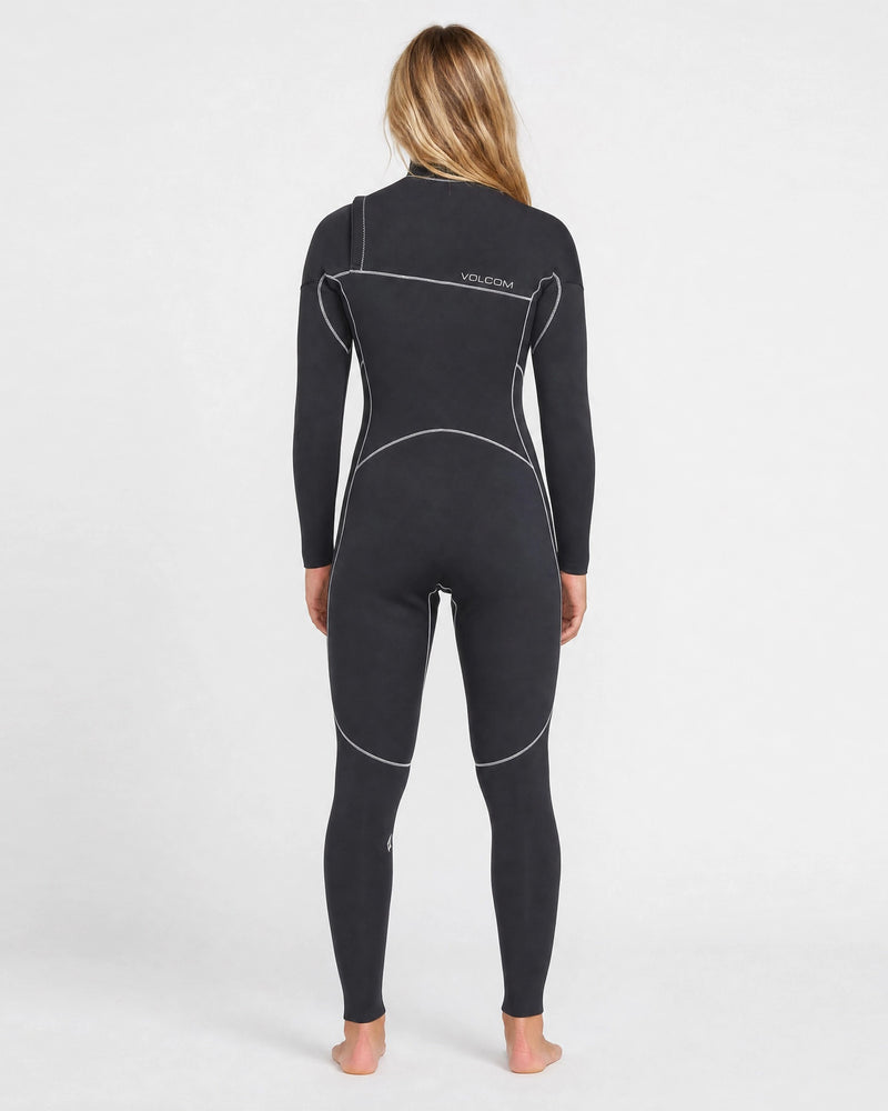 A person with long blonde hair stands barefoot, facing away, wearing a black Volcom Women's 3/2MM Modulator Chest Zip Full Wetsuit with white seams and “Volcom” on the back, against a plain white background.