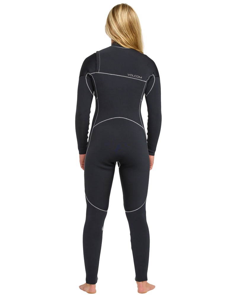 A person with long blonde hair stands barefoot, facing away, wearing a Volcom Women's 4/3 Modulator Chest Zip Full Wetsuit with white seams and the Volcom logo on the back.