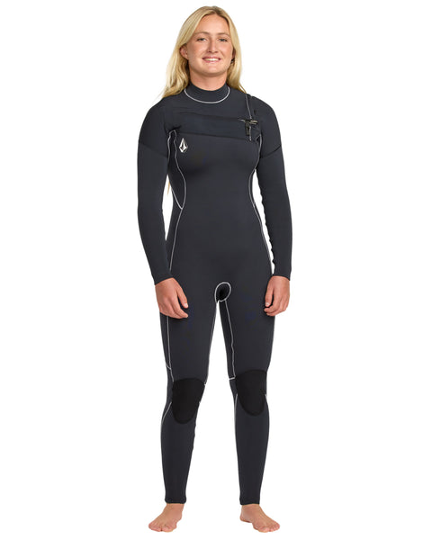 A woman with long blonde hair stands barefoot, smiling, and wearing the Volcom Women's 4/3 Modulator Chest Zip Full Wetsuit—a full-length black wetsuit with white seam accents and reinforced knee pads.