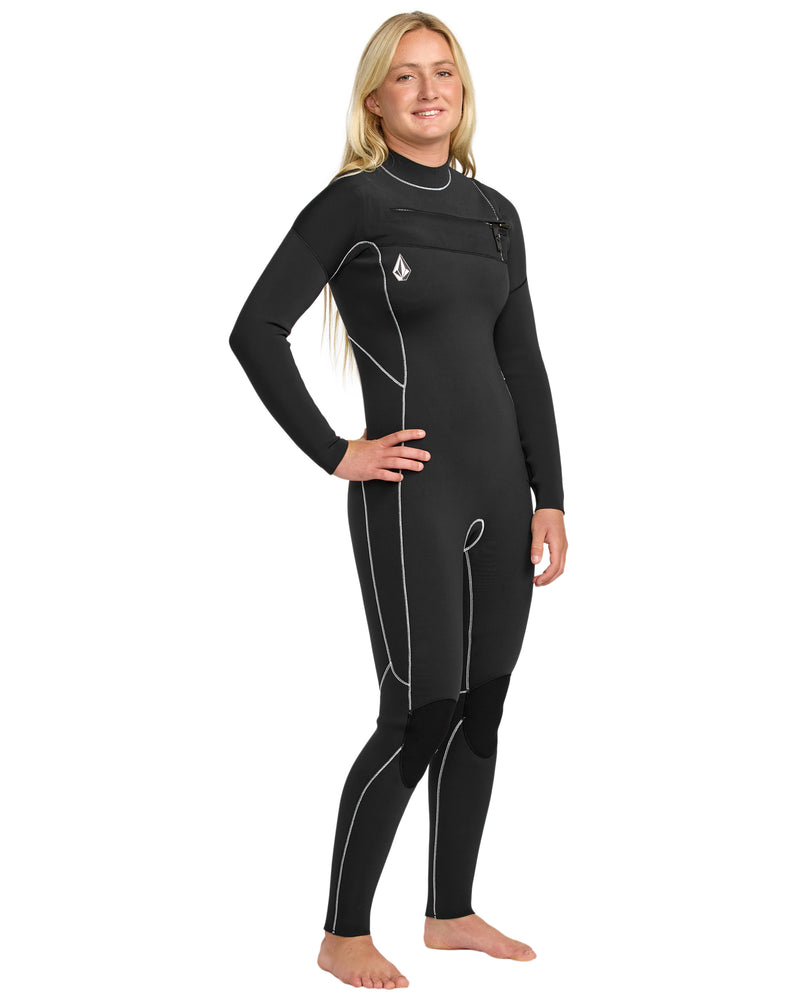 A woman with long blonde hair stands barefoot, smiling at the camera against a white background while wearing the Volcom Women's 4/3 Modulator Chest Zip Full Wetsuit in black with white seams.