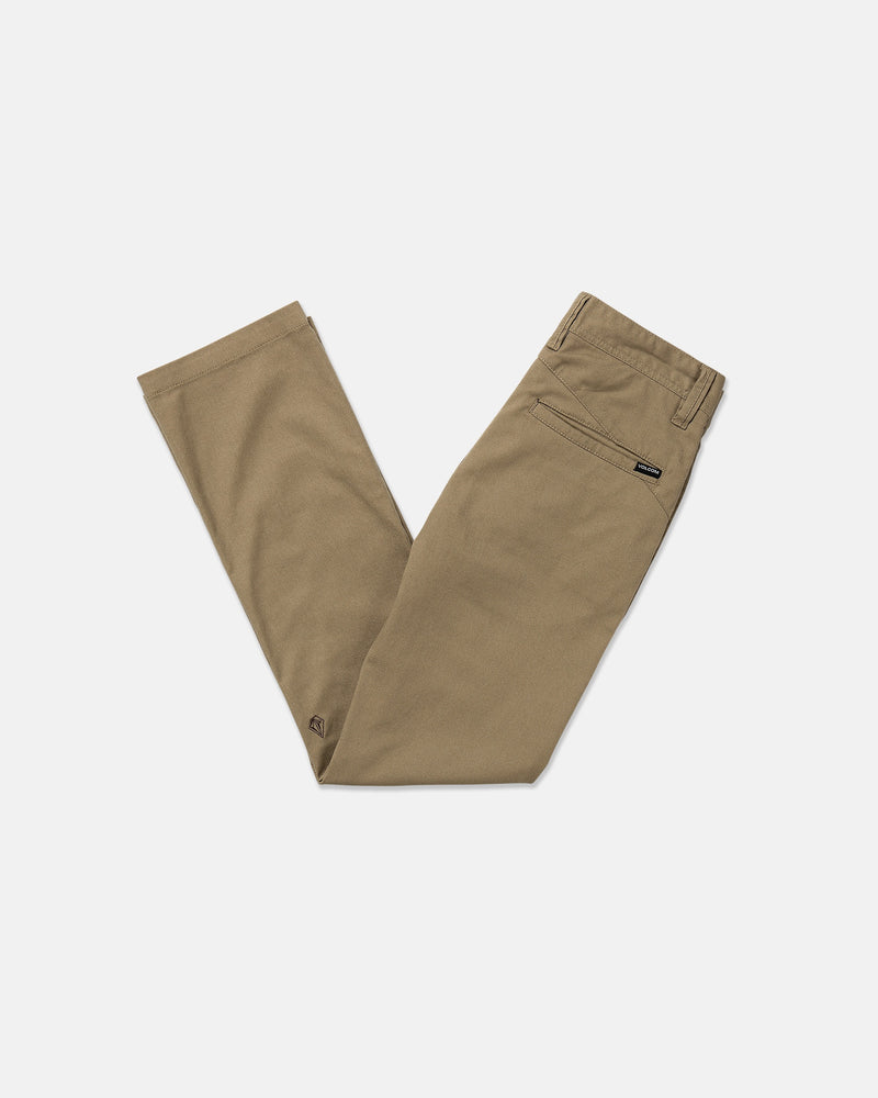 A pair of tan Volcom Kids' Big Boys' Frickin Modern Stretch Pants, featuring a back pocket and black brand tag, made with REPREVE recycled fibers, neatly folded with crossed legs on a plain white background.
