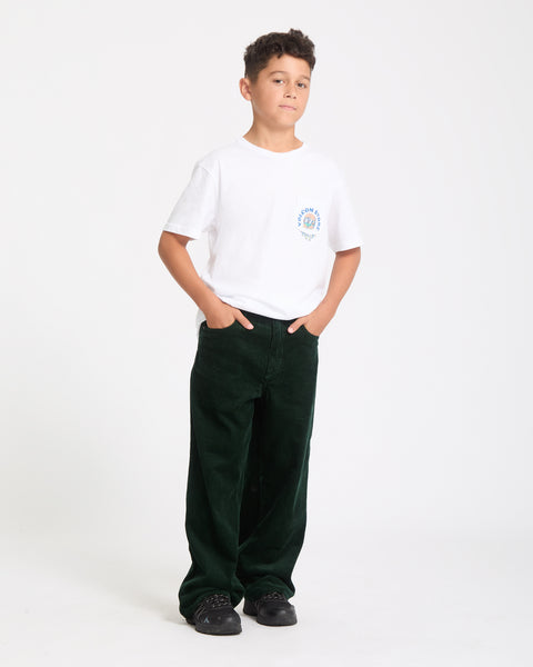 A boy stands against a plain white background with his hands in the pockets of his dark green corduroy pants, wearing a Kids' Burgstone Pocket Tee in organic cotton white and black sneakers.