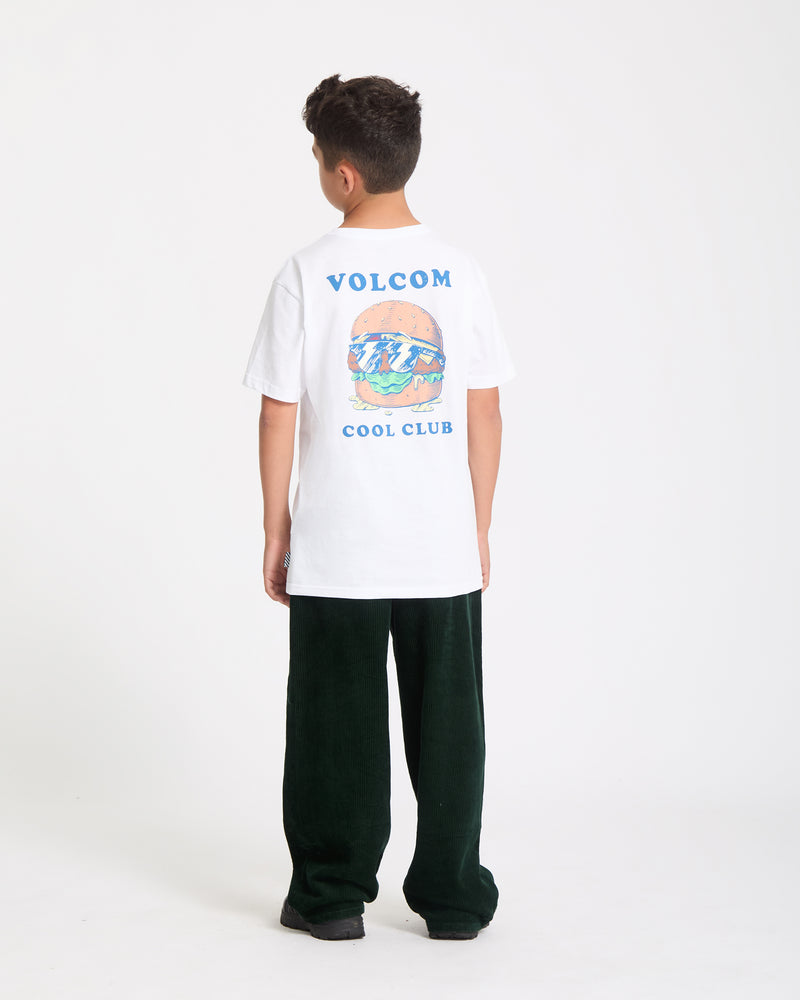 A boy faces away, wearing the Kids' Burgstone Pocket Tee—an organic cotton white T-shirt with burger and ocean art plus "VOLCOM COOL CLUB" text—paired with dark green pants and black shoes, set against a plain white background.
