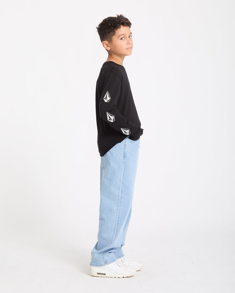 A boy stands in profile against a plain white background, wearing a Kids' Iconic Stone Gid Long Sleeve Tee with graphic sleeve patches, light blue jeans, and white sneakers.