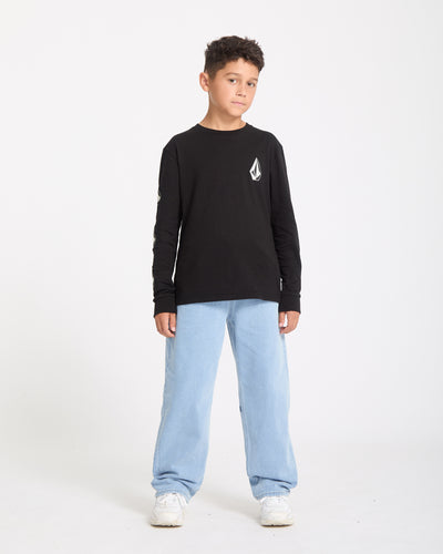 A boy stands against a white background, wearing the Kids' Iconic Stone Gid Long Sleeve Tee in black with a white chest emblem, light blue jeans, and white sneakers. He looks slightly to the side with a neutral expression.
