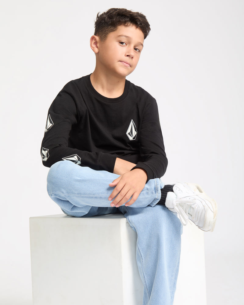 A boy with short brown hair sits on a white cube, confidently looking at the camera. He's wearing a black Kids' Iconic Stone Gid Long Sleeve Tee with white sleeve designs, light blue jeans, and white sneakers, one leg crossed.