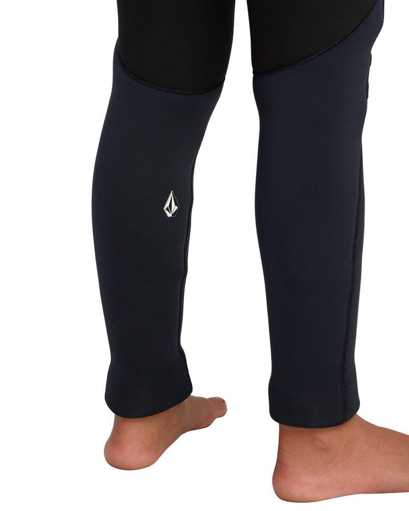 A close-up of the lower legs of a person standing barefoot, wearing the Volcom Kids' 5/4 mm Hooded Chest Zip Long Sleeve Full Wetsuit with a small white logo on one leg.