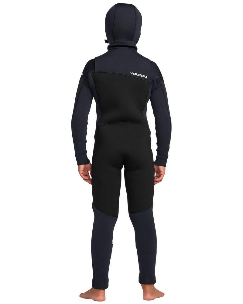 A child stands barefoot facing away in a black and dark blue Volcom Kids' 5/4mm Hooded Chest Zip Full Wetsuit, featuring the VOLCOM logo on the upper back and covering from head to ankles.