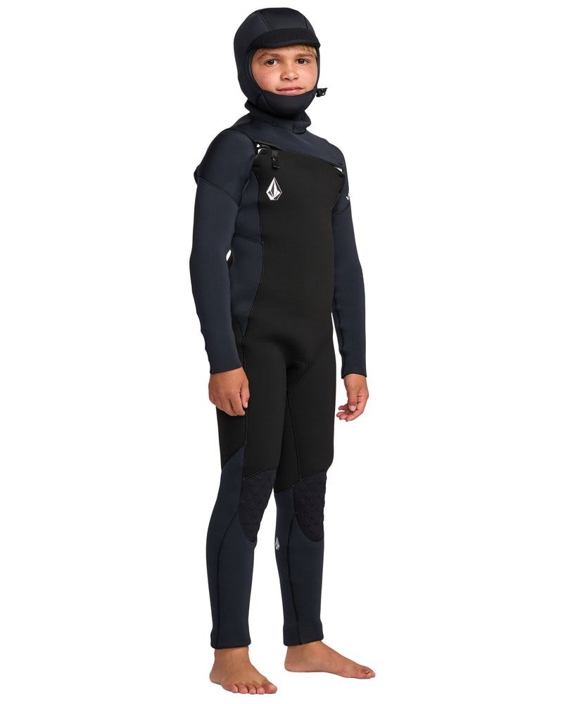 A young person stands barefoot against a white background wearing the Volcom Kids' 5/4 Mm Hooded Chest Zip Long Sleeve FullWetsuit, a black full-body wetsuit with an attached hood and chest logo.