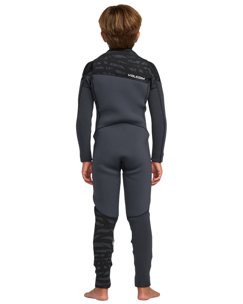 A young person with short brown hair stands barefoot, facing away, in a Volcom Kids' 3/2 Modulator Chest Zip Full Wetsuit. The wetsuit is black and dark gray with patterned details on the shoulders and lower left leg. Background is plain white.