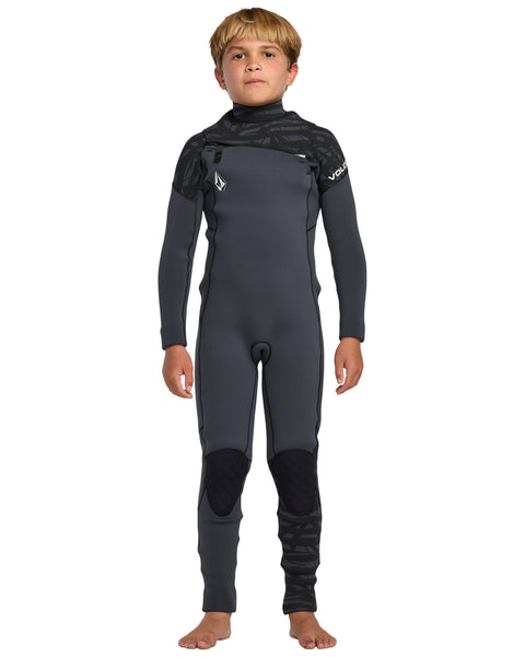 A young boy stands barefoot in the Volcom Kids' 3/2 Modulator Chest Zip Full Wetsuit—dark gray with black accents, reinforced knees, and a secure chest zip—posing confidently against a plain white background.