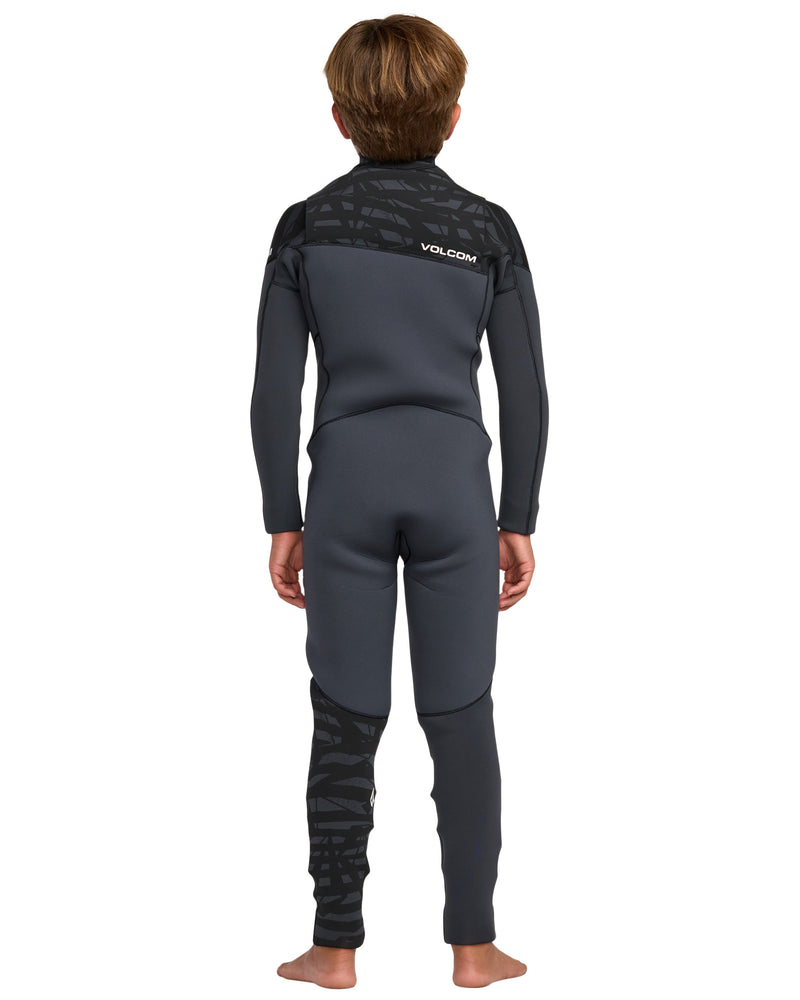 A child stands barefoot, facing away, wearing the Volcom Kids' 4/3 Modulator Chest Zip Full Wetsuit in black and dark gray with a textured pattern on the shoulders and lower left leg. The Volcom logo appears on the upper back.