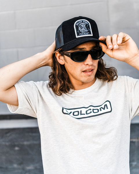 A person wearing a Volcom Men's Shred Supply Trucker Hat, black sunglasses, and a light-colored Volcom t-shirt stands outdoors, adjusting their hat with one hand. They have long brown hair and a neutral expression.