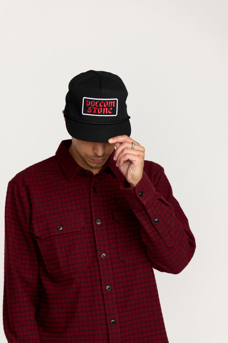 A person wearing a black Volcom Men’s Heavy Tone Hat and a red checked button-up shirt looks down while adjusting the brim of the cap with one hand.