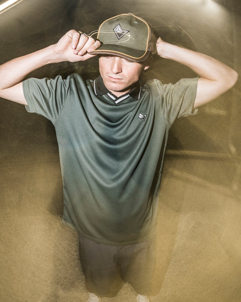 A person in a green shirt and hat stands outdoors at night, holding the brim of their Volcom Men’s V Line Adjustable Hat with both hands. The lighting creates a dreamy, blurred effect around them.