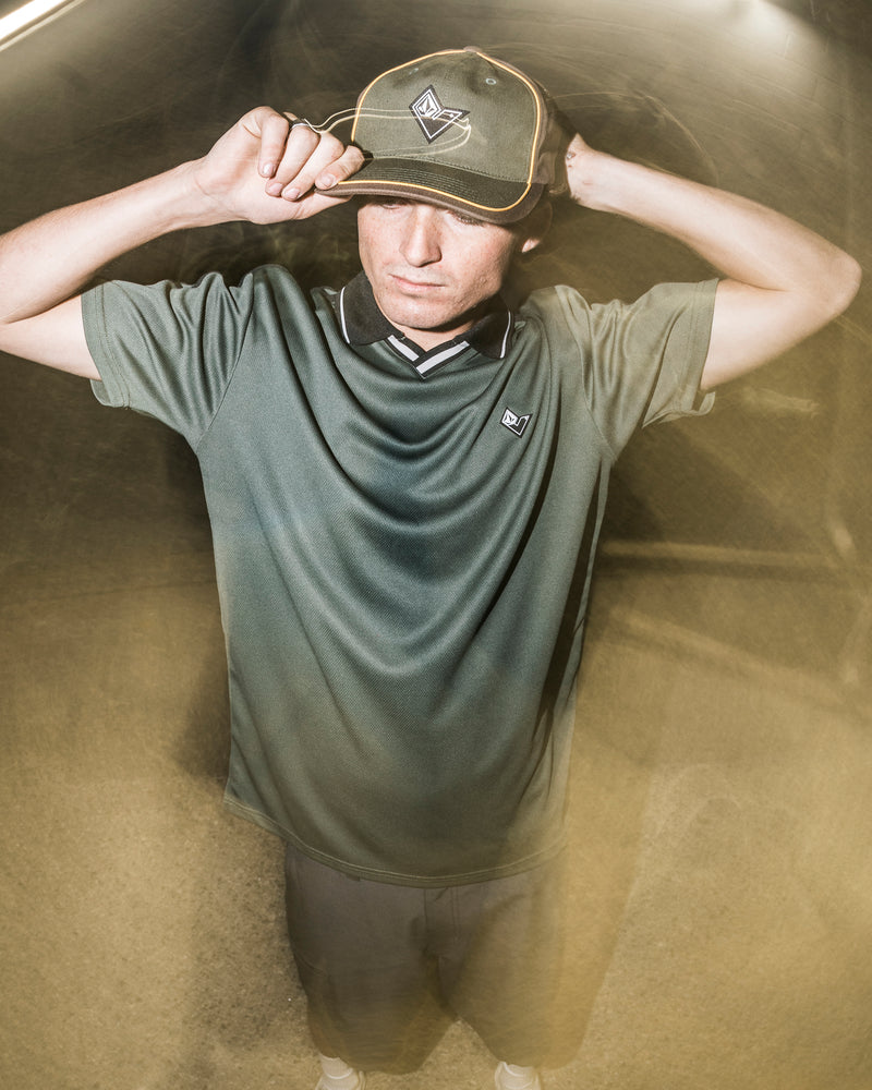 A person in a green shirt and hat stands outdoors at night, holding the brim of their Volcom Men’s V Line Adjustable Hat with both hands. The lighting creates a dreamy, blurred effect around them.