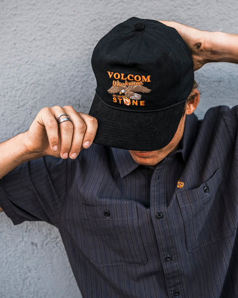 A man wearing the Volcom Men’s Stone Talon Workwear Hat, featuring a corduroy visor, and a striped shirt adjusts his cap while facing down against a gray wall background.