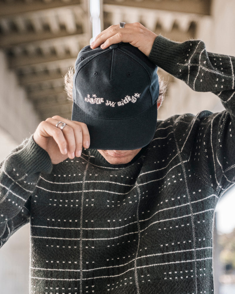 A person wearing a dark patterned sweater and a Volcom Men’s Alive We Ride Adjustable Hat with embroidered graphics pulls the cap down to partially cover their face while standing under an overhead structure.