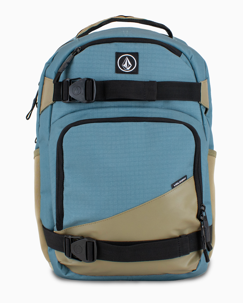 The Volcom Men's Grindstone Skate Backpack features a blue and tan design with black ergonomic straps, a top handle, front zippered pockets, a padded laptop sleeve, and a logo patch on the upper front.