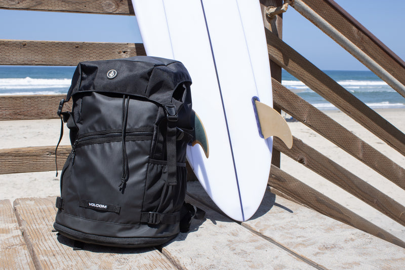 A Volcom Men's Litestone Gear Pack sits on wooden stairs beside a white surfboard with tan fins at the beach, showcasing durable construction and organized storage as waves and blue sky fill the background.