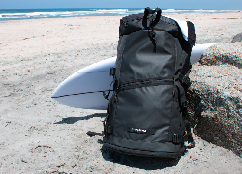 A Volcom Men's Litestone Gear Pack sits on a sandy beach near a large rock, with a white surfboard behind it. The durable backpack offers organized storage, set against the ocean waves under a clear sky.
