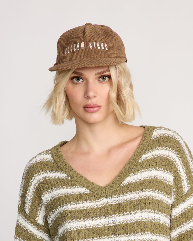 A woman with short blonde hair wears the Volcom Women's Full Stone Dad Hat in brown corduroy with white embroidery and an olive-green and white striped V-neck sweater, looking at the camera against a plain background.