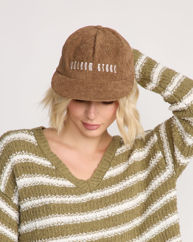 A person with short blonde hair wears a brown Volcom Women's Full Stone Dad Hat with "VOLCOM STONE" on the front and an olive-green striped cotton sweater. Their head is slightly down, one hand touching the cap's side.