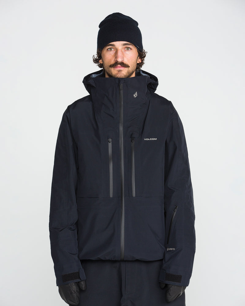 A man with a mustache wears a black beanie, black gloves, and the Volcom Men’s TDS® Inf Gore-Tex Jacket made from recycled nylon, standing against a plain white background.