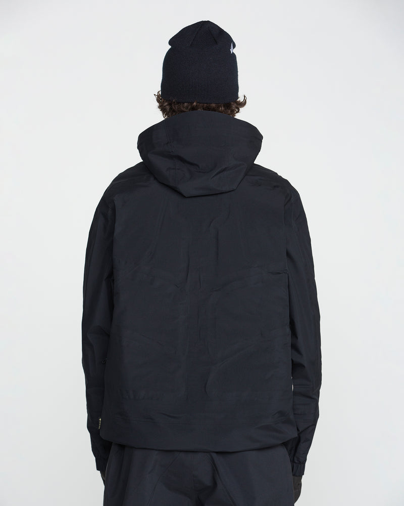 A person wearing a black Volcom Men’s TDS® Inf Gore-Tex Jacket with a hood and a black beanie stands facing away from the camera against a plain white background.