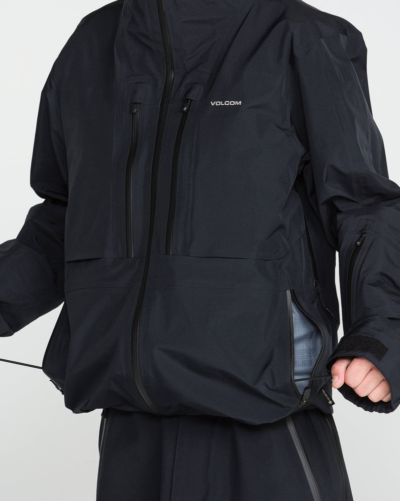 A person is shown from shoulders to waist wearing the Volcom Men’s TDS® Inf Gore-Tex Jacket in black. The GORE-TEX jacket features multiple zippered pockets and an adjustable drawstring hem, which the person is pulling. Their face and lower legs are not visible.