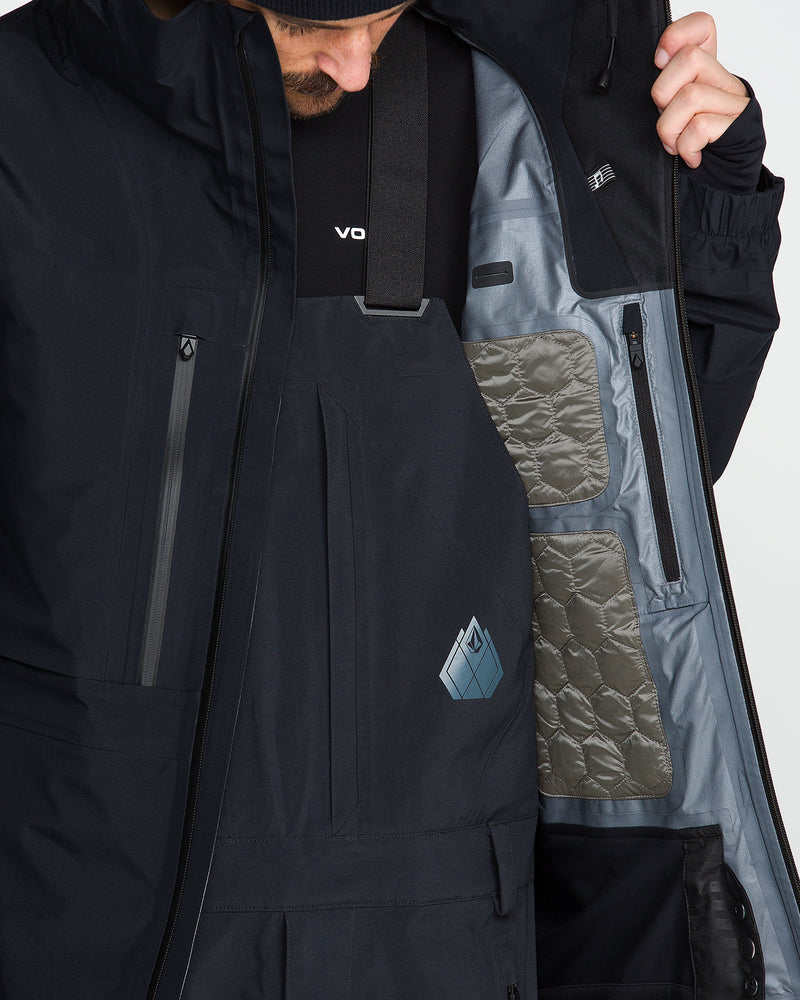 Close-up of a person wearing a dark ski jacket and bib pants, opening the jacket to show the light gray interior lining with quilted pockets.