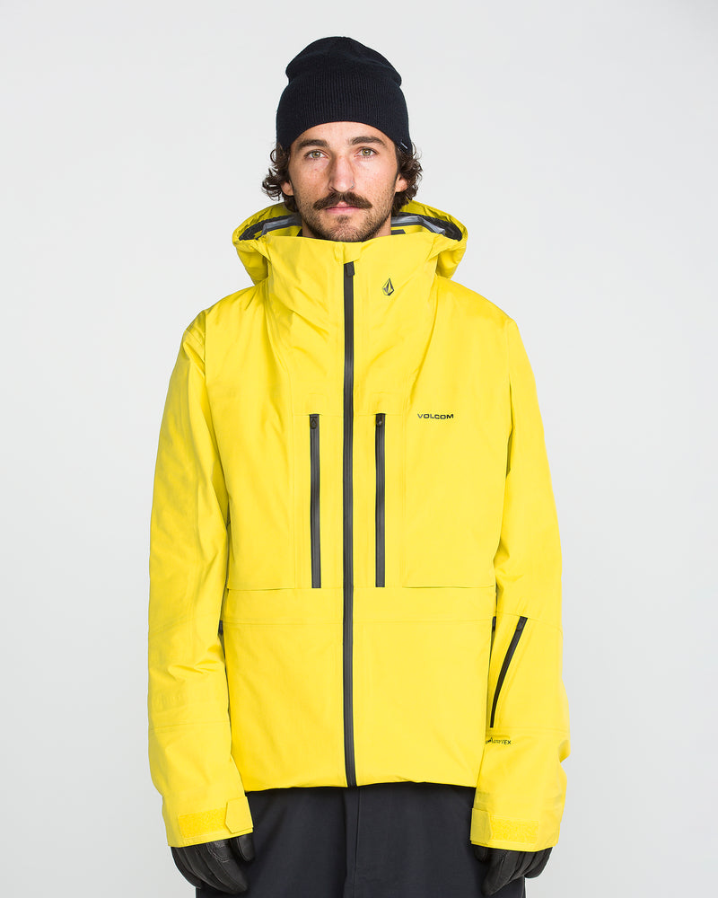 A person wearing the Volcom Men’s TDS® Inf Gore-Tex Jacket in yellow, along with black gloves, a black beanie, and dark pants, stands facing the camera against a plain light background.