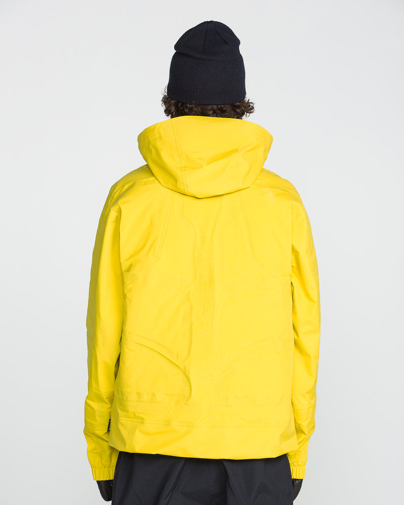 A person wearing the Volcom Men’s TDS® Inf Gore-Tex Jacket in yellow, paired with a black beanie and black pants, is standing with their back to the camera against a plain light background.