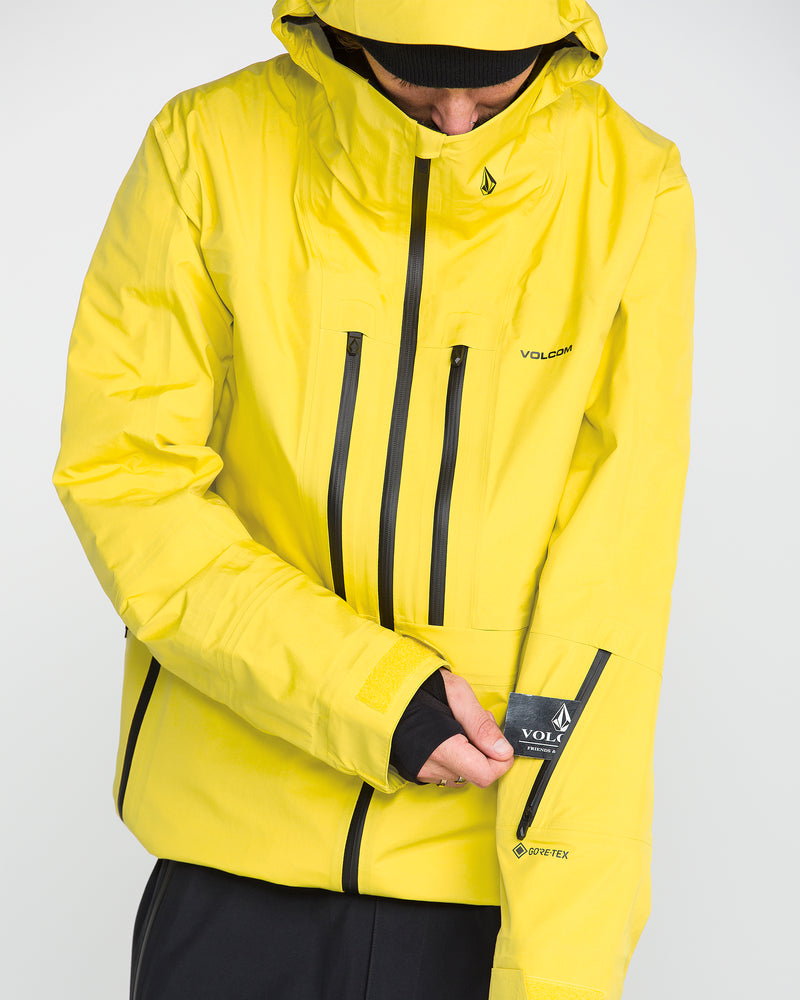A person wears the Volcom Men’s TDS® Inf GORE-TEX Jacket, its bright yellow color and black zippers standing out as they pull an item from a zippered sleeve pocket. The recycled nylon jacket features the hood up, partially obscuring their face.