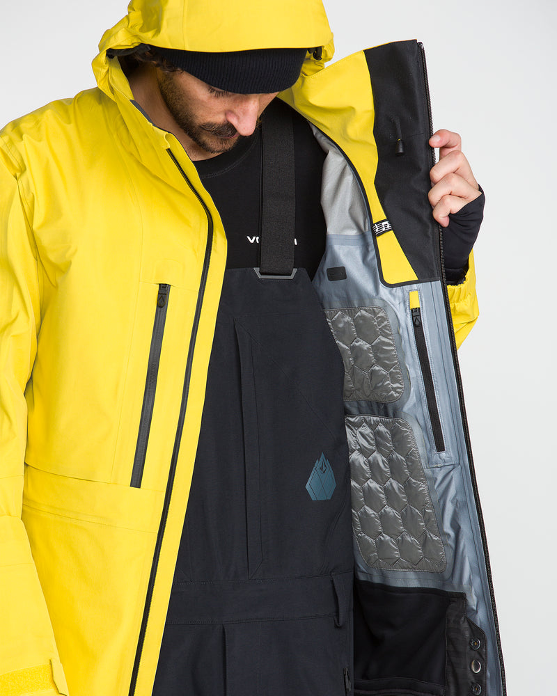 A person wearing a yellow Volcom Men’s TDS® Inf Gore-Tex Jacket opens it to reveal a quilted inner lining and black overalls underneath. The jacket’s hood partially obscures their face as they look down.